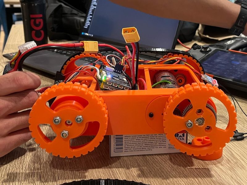 An early orange 3D-printed chassis with geared wheels, held in one hand